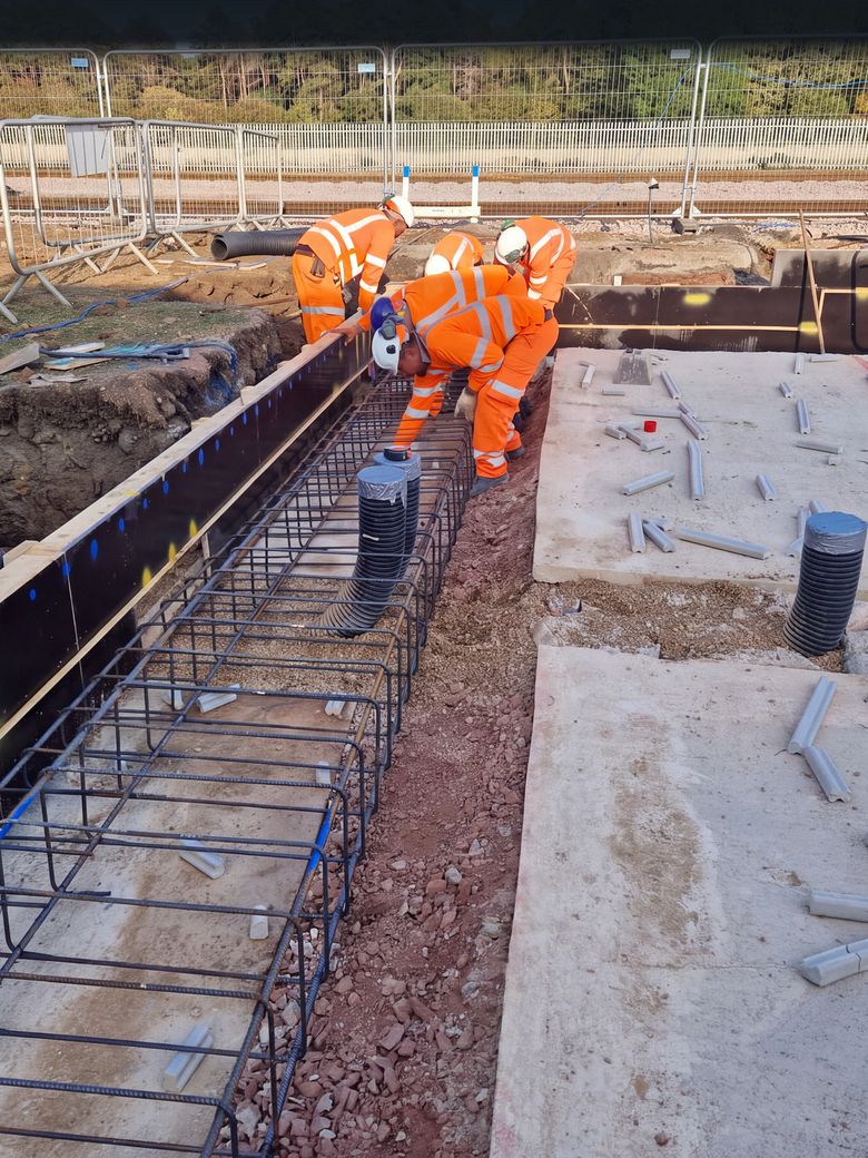 Groundworkers in high visibility clothing lowering a ground beam reinforcement cage into timber shuttering
